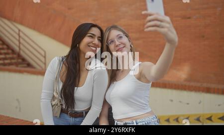 Two women smiling and taking a selfie together on a city street with exposed brick architecture in the background Stock Photo