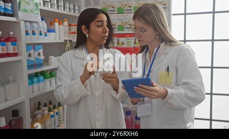 Two women pharmacists in lab coats discuss a medication in a drugstore filled with various health products. Stock Photo