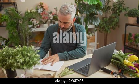 Handsome bearded man taking notes in a flower shop, surrounded by greenery and blooms, with a laptop on a table. Stock Photo
