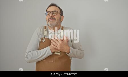 Middle-aged bearded man in glasses and apron over isolated white background expresses heartfelt gratitude with hands on chest. Stock Photo