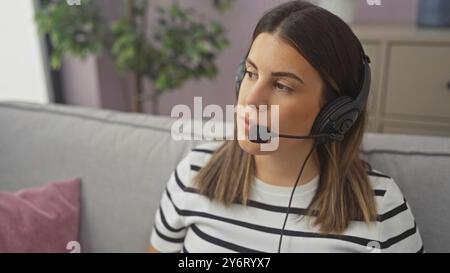 A focused young woman with a headset works remotely from her cozy living room couch. Stock Photo