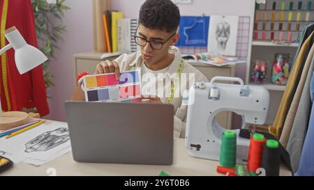 A young man compares fabric colors during a video call in a tailor shop surrounded by sewing equipment. Stock Photo