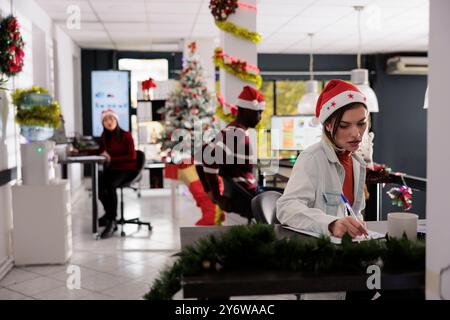 Multiethnical business project team wearing christmas hats working in festive office. Hard working employees doing tasks and comparing graphs in decorated workplace during christmas season Stock Photo