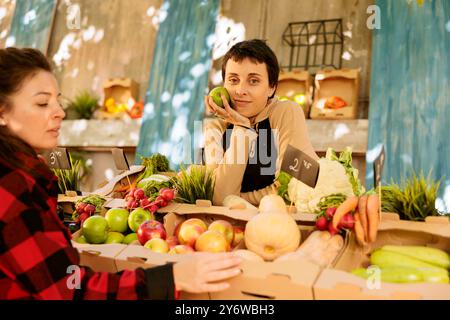 Female customer selecting fruits and vegetables while vendor stands behind counter with an apple in hand. Portrait of woman farmer selling variety of seasonal organic fresh produce at harvest fair. Stock Photo