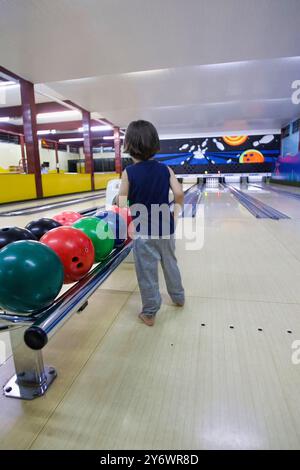 A young boy is standing in front of a bowling lane with a ball in his hand. The bowling balls are lined up on the lane, and the boy is about to throw Stock Photo