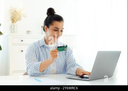 Transferring money, e-banking, online shopping. Beautiful happy indian or arabian young woman holding a credit card and using a laptop for making a purchase on internet while sitting at a desk, smiles Stock Photo