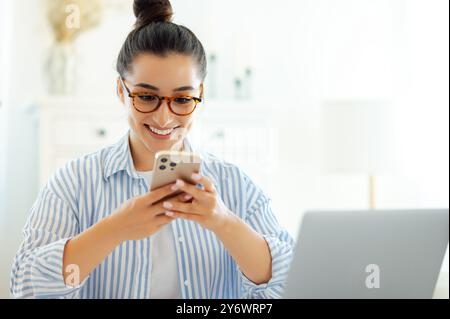 Joyful excited indian or arabian young woman with glasses, using her mobile phone, chatting online with clients or colleagues about work issues while sits at a workplace, smiles. Gadgets and devices Stock Photo