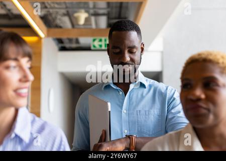Holding laptop, african american man checking time on watch while standing with colleagues Stock Photo