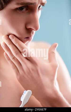 Young man is holding a toothbrush and touching his face with a thoughtful expression. He may be contemplating his oral hygiene routine or experiencing Stock Photo