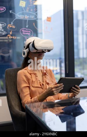 Using VR headset, woman holding tablet and sitting in modern office Stock Photo