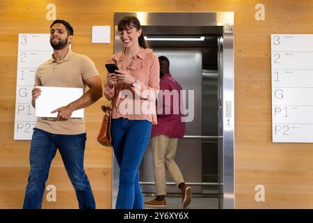 Walking in office, man holding laptop while woman using smartphone, smiling Stock Photo