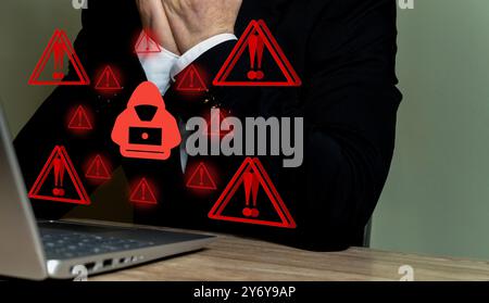 A businessman in a suit sitting at a desk with a laptop, surrounded by red warning symbols and a hacker icon. Stock Photo