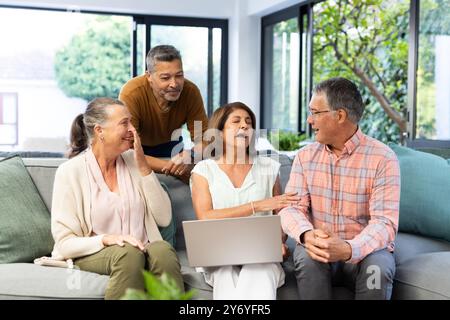 Using laptop, senior diverse friends sitting on couch and talking, enjoying time together, at home Stock Photo