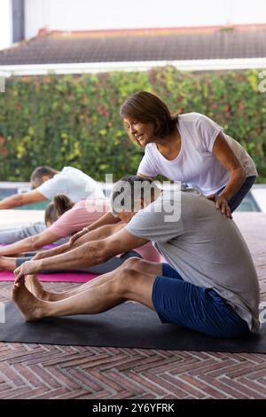 Assisting senior man stretching on yoga mat during outdoor fitness class, at home Stock Photo