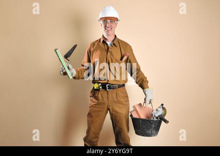 Construction worker dressed in work clothes holding tools and building materials isolated on brown background. Front view. Stock Photo