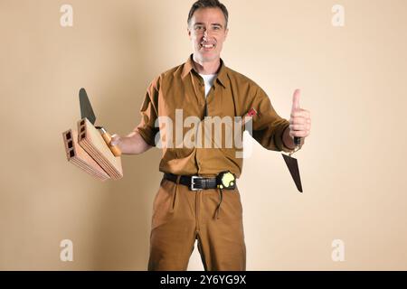 Smiling bricklayer making ok gesture with finger holding construction tools and materials and dressed in work clothes isolated on brown background. Fr Stock Photo