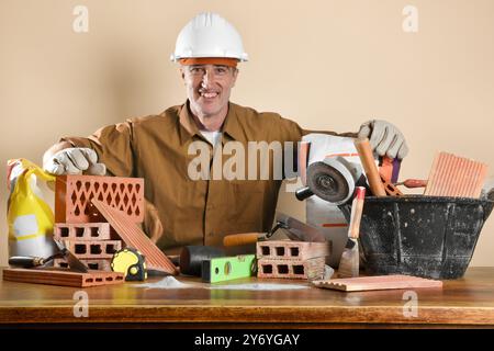Smiling worker dressed in protective clothing and gear standing behind a wooden table showing construction materials and tools. Front view. Stock Photo