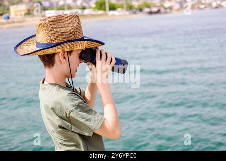A boy in a straw hat is looking through binoculars at the sea Stock Photo