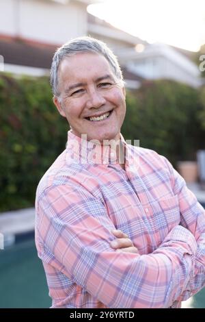 Smiling senior man in plaid shirt standing with arms crossed outdoors, at home Stock Photo