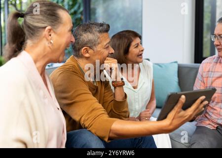 Laughing together, senior diverse friends using tablet and enjoying conversation on couch, at home Stock Photo
