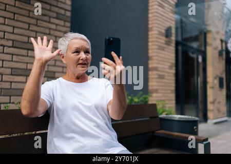 Senior woman sitting on urban bench outdoors, waving and talking on video call on smartphone. European gray-haired older female dressed casually Stock Photo