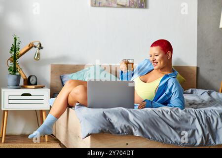 In a cozy bedroom, a young woman sips tea, lounging on her bed with a laptop open. Stock Photo