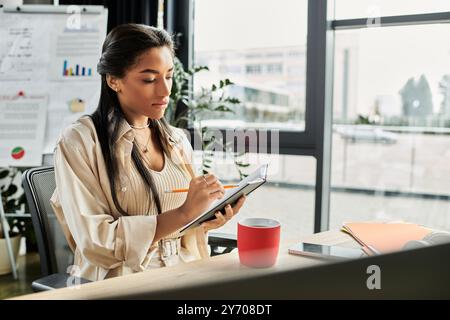 A focused young woman writes notes while sitting at her office desk with large windows. Stock Photo