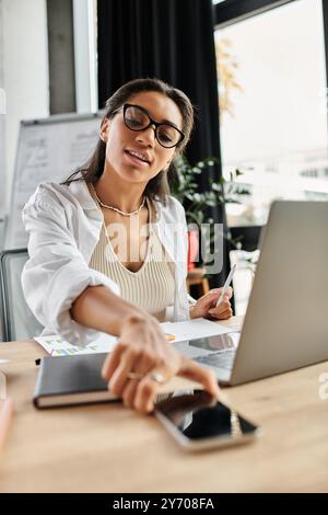 A focused young woman is analyzing data on her laptop while reaching for her phone. Stock Photo