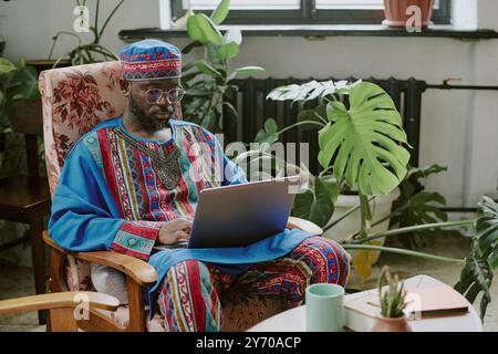 Mature African American man dressed in traditional costume sitting in armchair and working in modern biophilic office Stock Photo