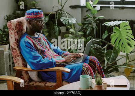 Side view shot of African American man in traditional costume typing on keyboard while working in cozy office Stock Photo