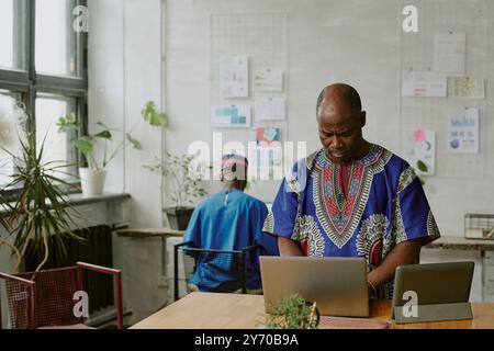 Mature African American programmer using tablet and laptop while hi African African colleague sitting at work table in background Stock Photo