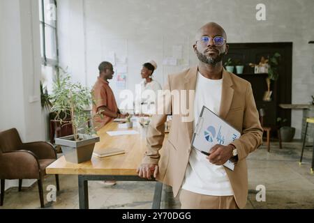 African American confident businessman leaning on table while posing for photo, his partners chatting in blurred background Stock Photo