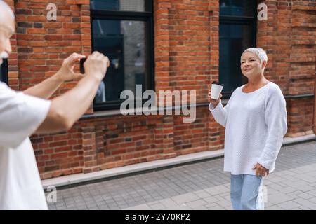 Gray-haired retired male photographer happily taking photo on smartphone of senior wife holding coffee cup outside charming brick building, exploring Stock Photo