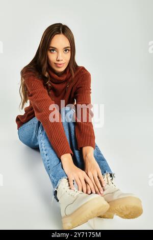 A beautiful young woman sits on the floor, dressed in a stylish sweater and jeans, radiating warmth. Stock Photo