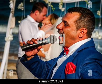 Wearing a red bow tie and matching pocket square, the master of ceremonies calls into the microphone as the bride and groom share their wedding kiss on the beach of the Black Sea in the background. Taken at a Bulgarian beach wedding in Albena, Balchik Municipality, Bulgaria. Stock Photo