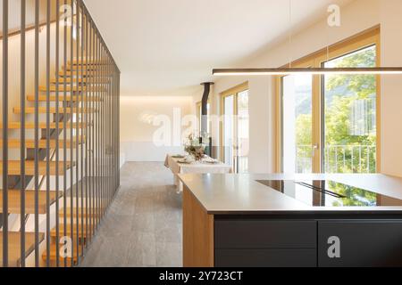 Frontal view of open space with a kitchen island, a table and at the back the sofa and the stove. On the left a modern staircase in wood and metal. No Stock Photo