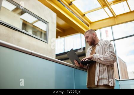 Side view of adult man wearing casual clothes focused on using laptop while standing on business center balcony Stock Photo