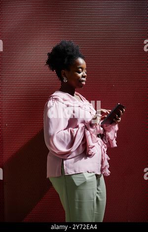 Vertical shot of smiling African American businesswoman standing in hard light against red wire grid using digital tablet for work in office center, copy space Stock Photo