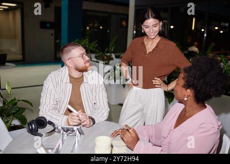 Diverse creative team of two colleagues sitting at cafe table participating in business meeting with supportive female boss checking up on work progress at office center, camera flash Stock Photo