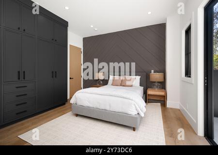 Modern bedroom with dark built-in wardrobes, a cozy bed, and wooden side tables, featuring a chevron-patterned accent wall and natural light. Stock Photo
