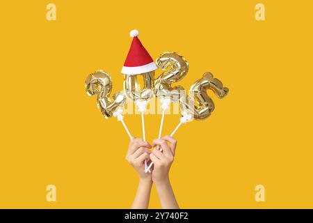 Female hands with figure 2025 made of foil balloons and Santa hat on yellow background Stock Photo