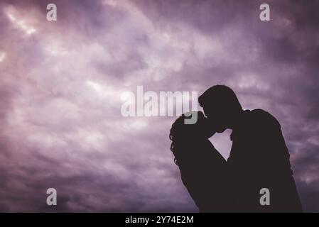 Photography of silhouetted couple embracing in a kiss set against a purple storm clouded sky at dusk Stock Photo