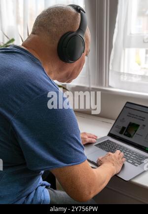 A senior man at home with a wireless headphone and a laptop computer, learning and try to keep up to date with modern technology. A technology use con Stock Photo