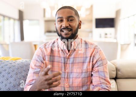 Talking and gesturing, smiling indian man in casual shirt sitting on couch, on video call Stock Photo