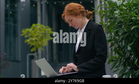 Smiling Caucasian girl female woman businesswoman student worker freelancer sit outdoors holding coffee cup sitting working typing online laptop pc Stock Photo