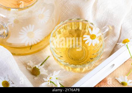 Close up glass transparent cup full of healthy chamomile tea, glass teapot, daisies on white napkin on wooden tray. Tea drinking for health. Chamomile Stock Photo