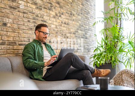 Handsome confident developer working on software over laptop while sitting on sofa in modern office Stock Photo