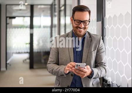 Successful male entrepreneur smiling and checking messages over mobile phone in office corridor Stock Photo