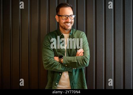 Male entrepreneur with arms crossed smiling and contemplating while standing over black wall in office Stock Photo