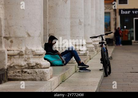 Vicenza, Italy - September 13, 2024: A Deliveroo worker takes a break sitting on the stairs, leaning against his work bag with his bicycle resting aga Stock Photo